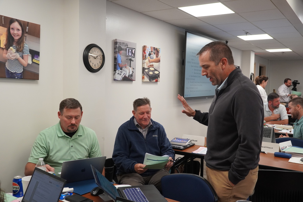 Three school administrators, two sitting and one standing, are chatting together and working collaboratively. 