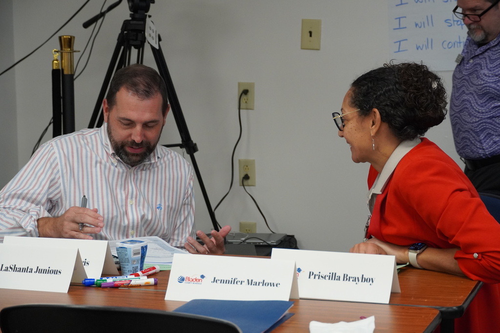 Two colleagues sit facing each other at a table smiling and speaking to one another. 