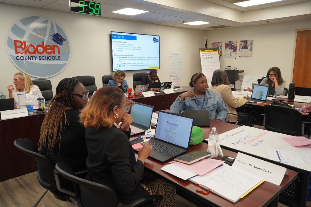 Adults sit together, working collaboratively at tables with computers in front of them.