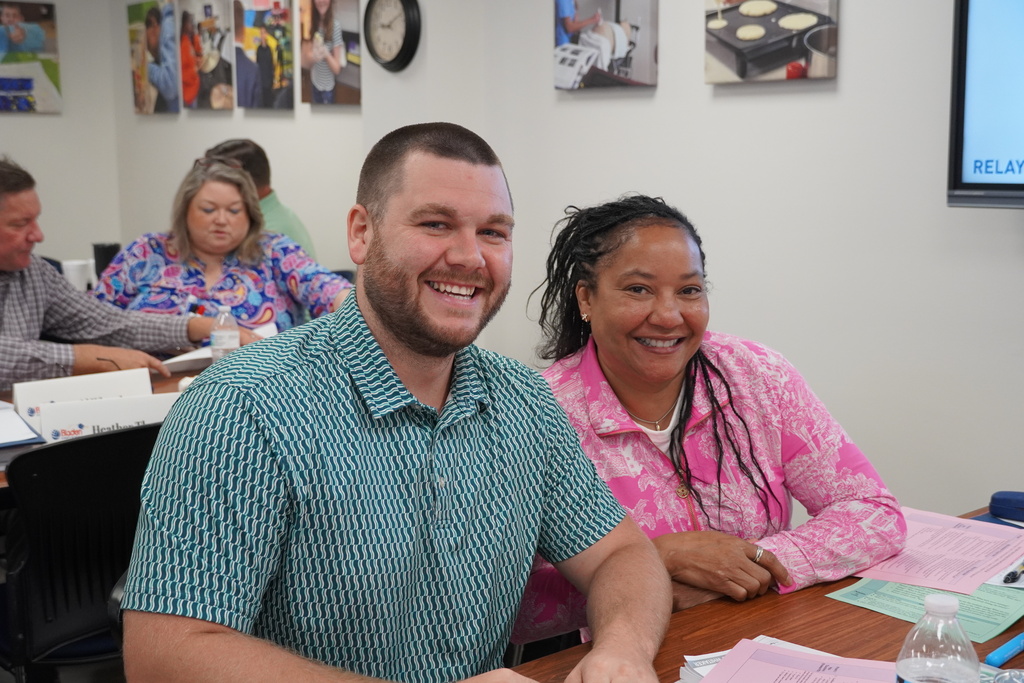 To school administrators sit smiling together at a desk. Behind them are more school administrators working together. 