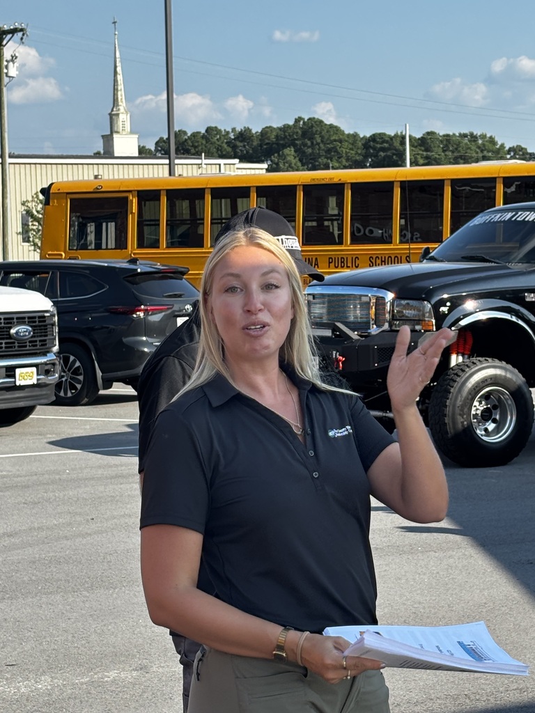 An instructor stands in a parking lot with a school bus behind her, speaking.