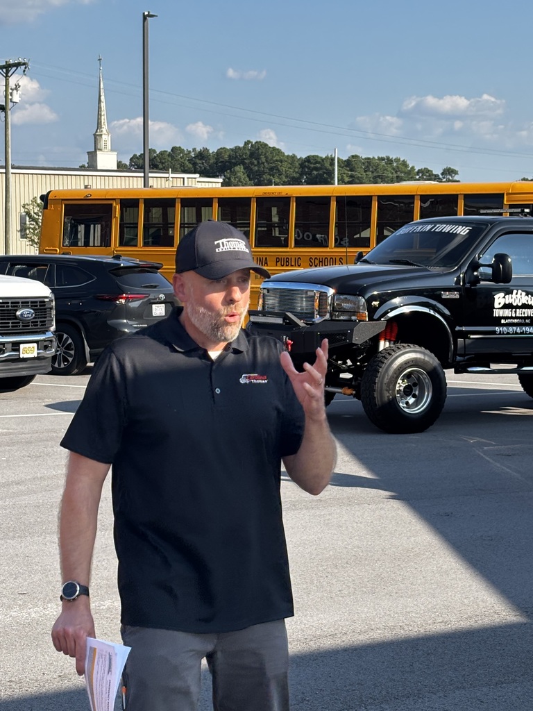 An instructor is speaking outside in a parking lot. Behind him are vehicles, including a school bus.