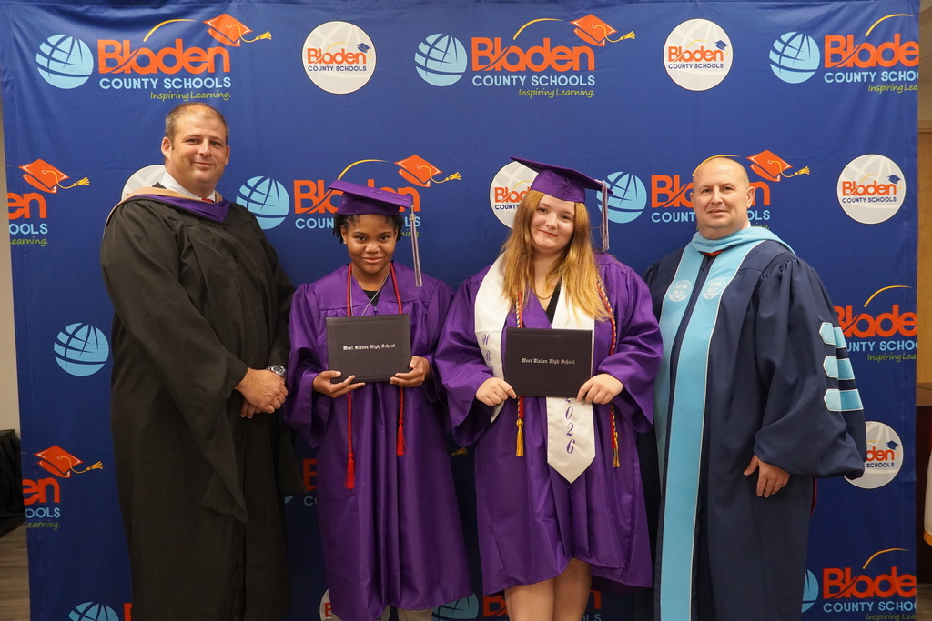 Two administrators wearing ceremonial robes stand beside two graduates wearing purple caps and gowns smiling. The graduates are holding their diplomas.