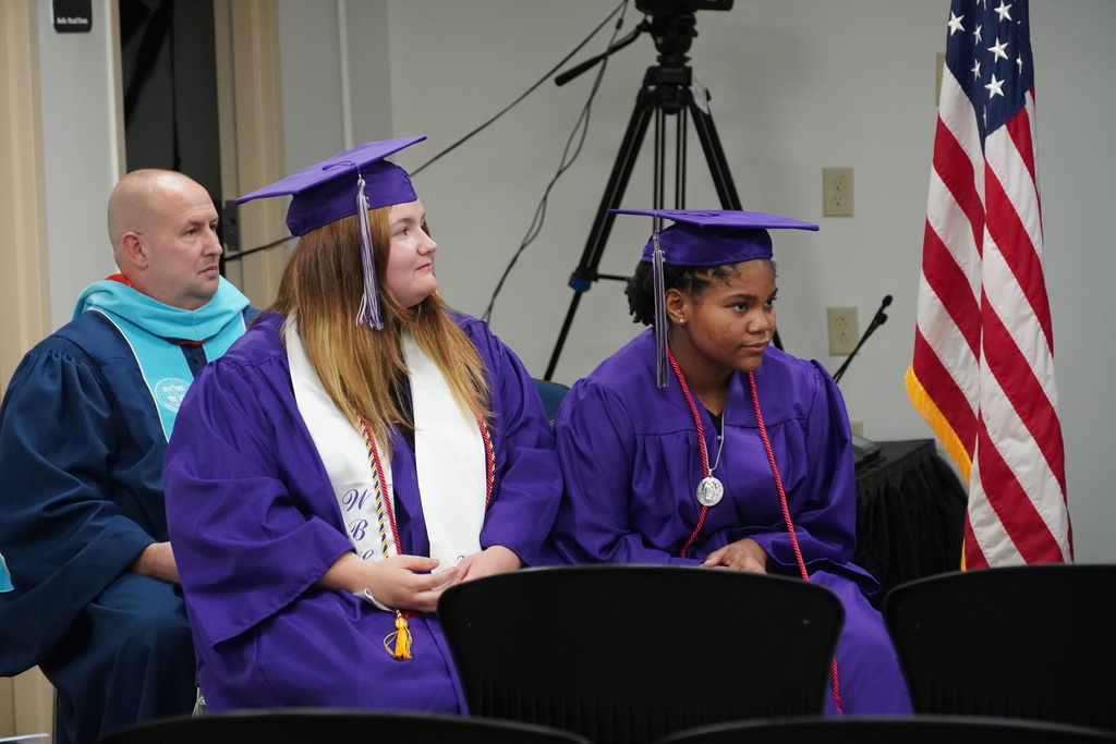 Two high school graduates wearing purple robes and caps are seen seeting next to the American Flag. Behind them is the Bladen County Schools superintendent.