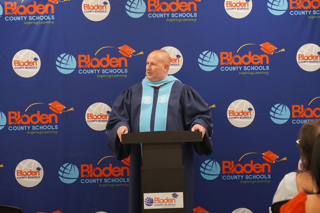 An adult is wearing a ceremonial robe for graduation and speaking from a podium in front of a Bladen County Schools step and repeat banner.
