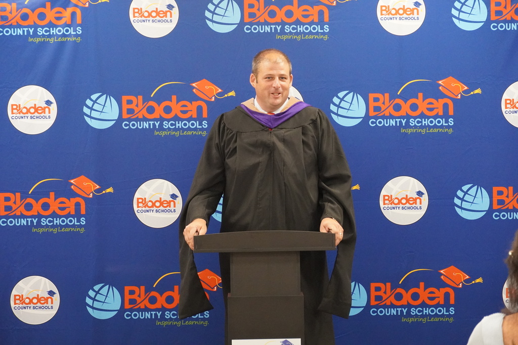 West Bladen Principal Chris Orrock stands at a podium wearing a graduation robe. Behind him is a Bladen County Schools step and repeat banner.