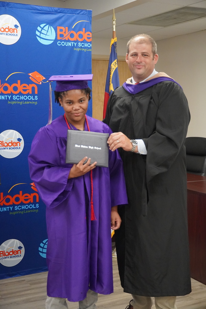 A graduate wearing a purple gown and cap stands smiling with a school administrator while holding a diploma.