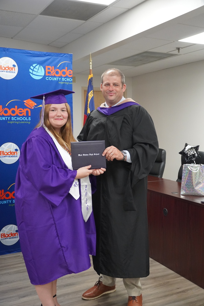 A graduate wearing a purple gown and cap stands smiling with a school administrator while holding a diploma.