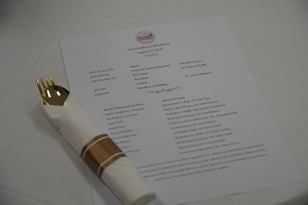A printed agenda titled New Employee Breakfast sits on a table with wrapped plastic silverware. 