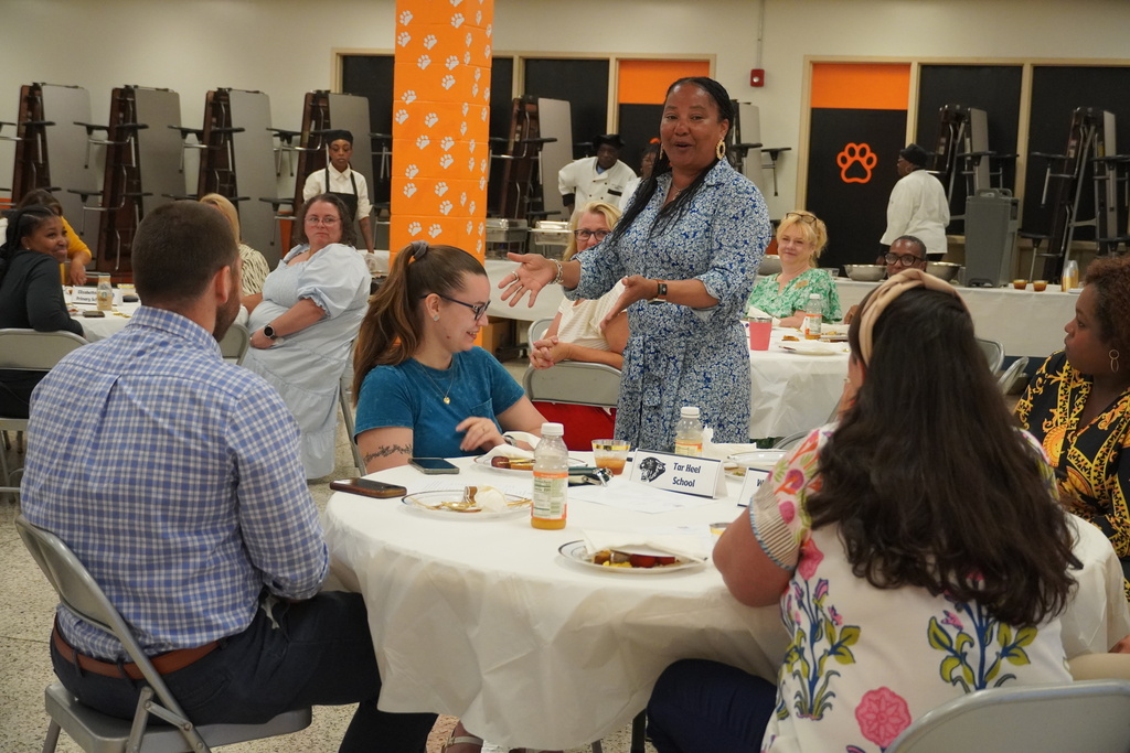An adult is standing up introducing a new employee who is seated next to her.