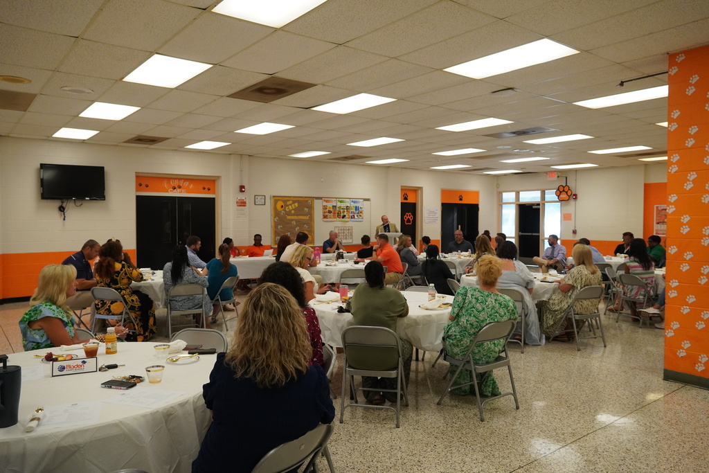 A group of adults sit in a school cafeteria listening to someone speak from the podium. 