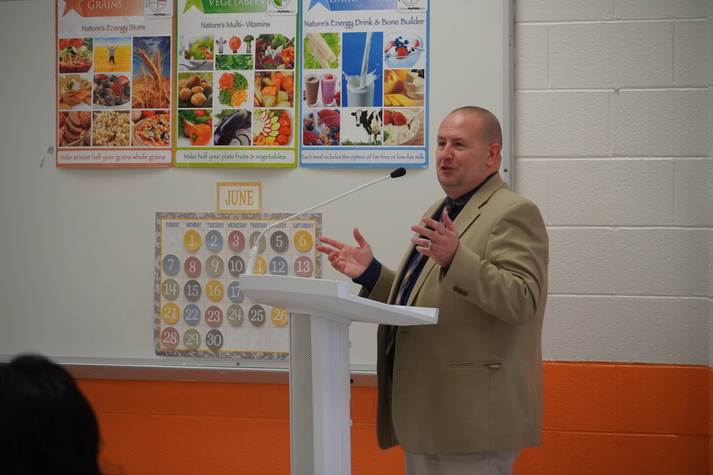 The superintendent is standing at a podium smiling and speaking to a crowd.