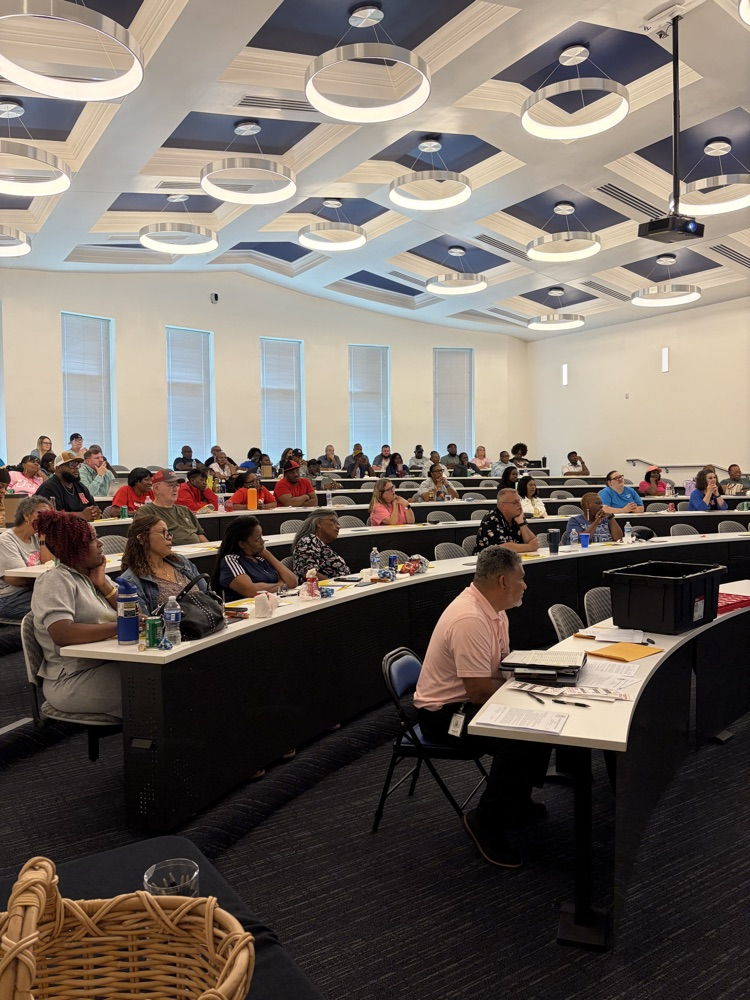 Adults sit in an auditorium class behind desks listening to a presenter.