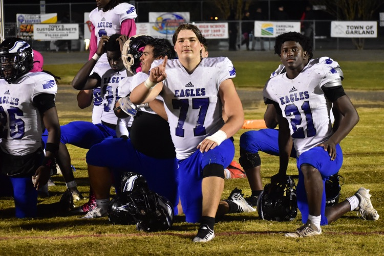 football players kneeled on the ground wearing blue bottoms white jerseys with blue numbers player in the front is holding up the number one sign by the rest of the team talks