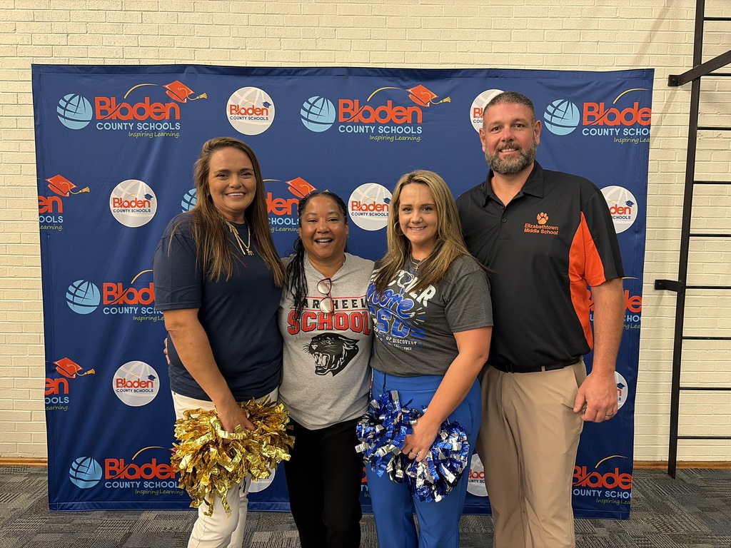 Four administrators stand together smiling in front of the Bladen County Schools step  and repeat banner.