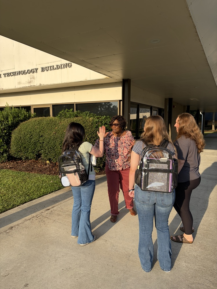Two students and two adults greet each other outside of a school building. The students are wearing clear backpacks.