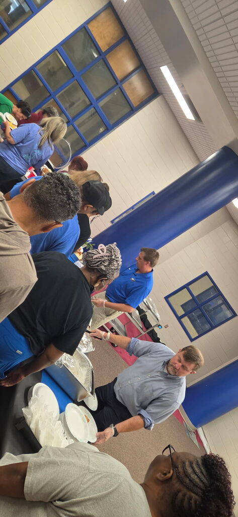 5 adults stand in line waiting to be served lunch, a man in a light blue dress shirt and a man in a royal blue shirt are serving food. They are in a room that has white walls with blue columns in the background.