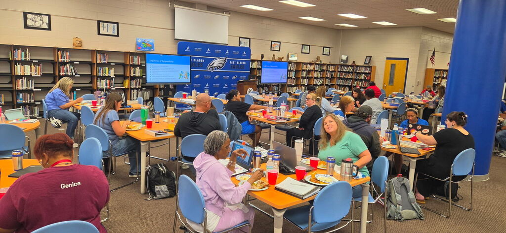 Adults in a library at tables, eating lunch. There is a blue column on the right, tvs in the back, an East Bladen backdrop, and bookshelves with books. 