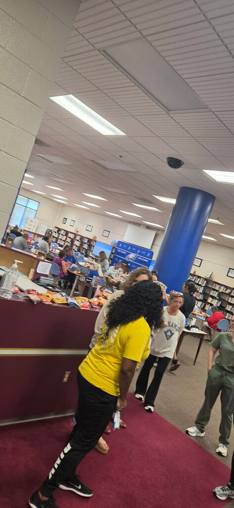 5 adults stand in line waiting to be served lunch, there is a lady in a yellow shirt at the front. They are in a library that has white walls. burgandy carpet, with blue columns in the background.