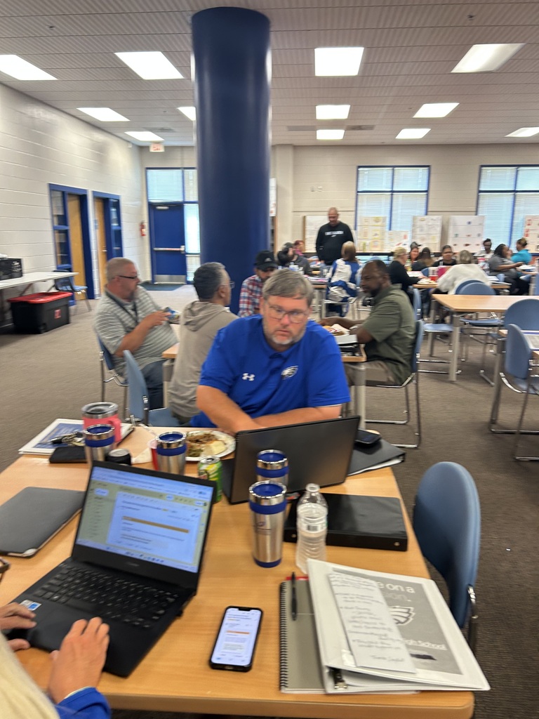 Adults in a library at tables, eating lunch. There are tvs in the back, an East Bladen backdrop, and bookshelves with books. 