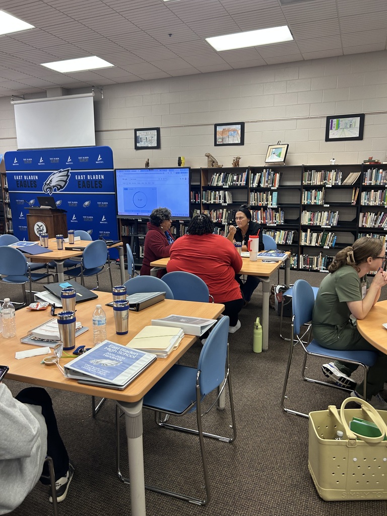 Adults in a library at tables, eating lunch. There are tvs in the back, an East Bladen backdrop, and bookshelves with books. 