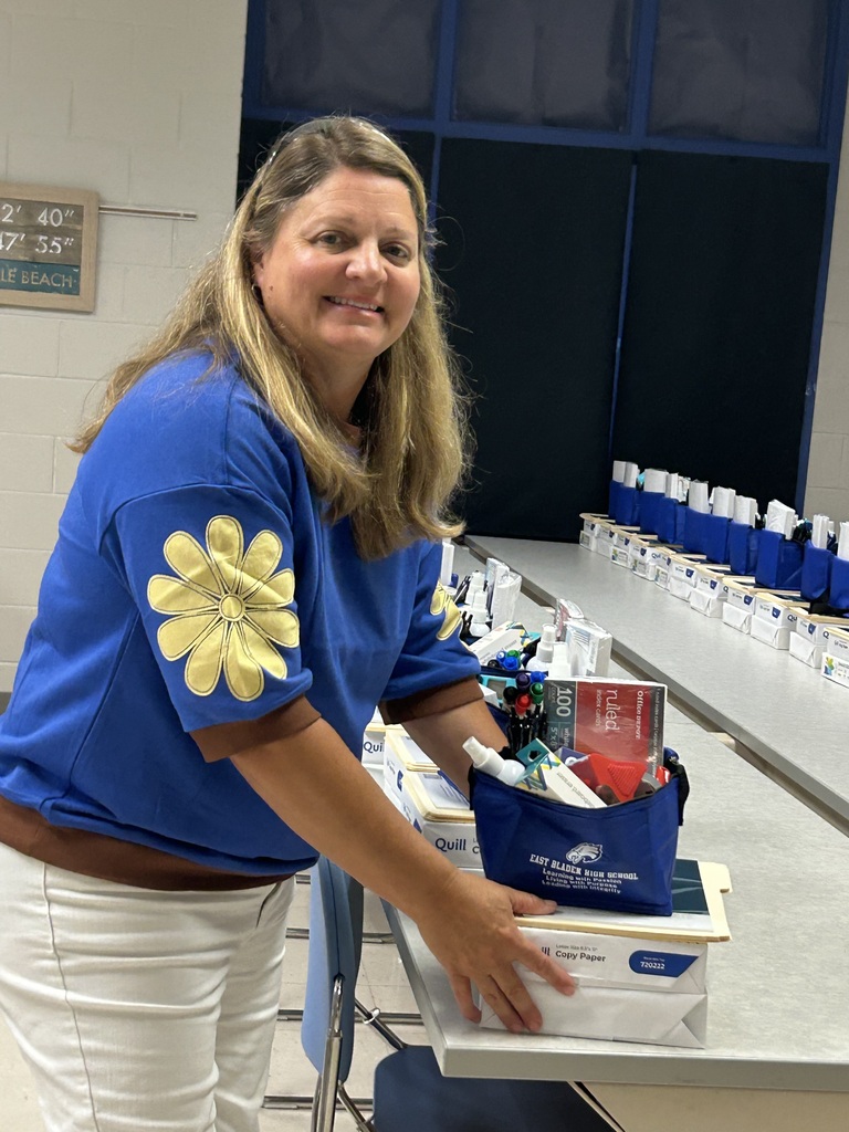 Lady in blue shirt with a yellow flower on it smiles for the camera and she picks up her gift of school supplies and a East Bladen lunch box.