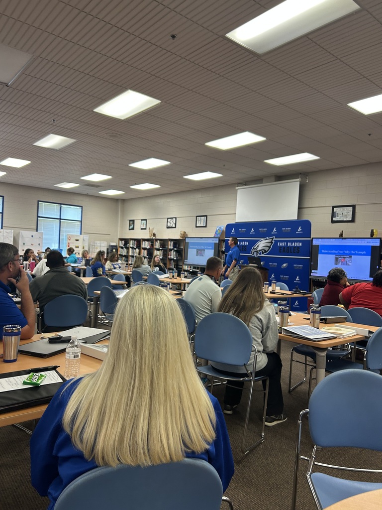Adults in a library at tables, eating lunch. There are tvs in the back, an East Bladen backdrop, and bookshelves with books. 