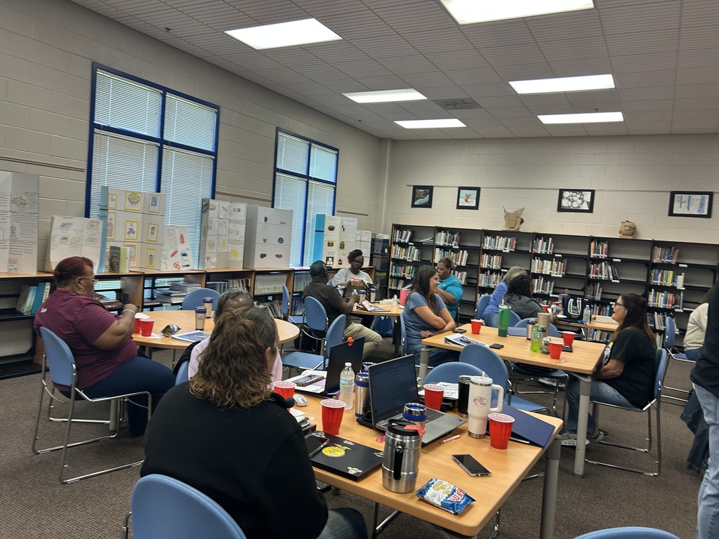 Adults in a library at tables, eating lunch. There are tvs in the back, an East Bladen backdrop, and bookshelves with books. 