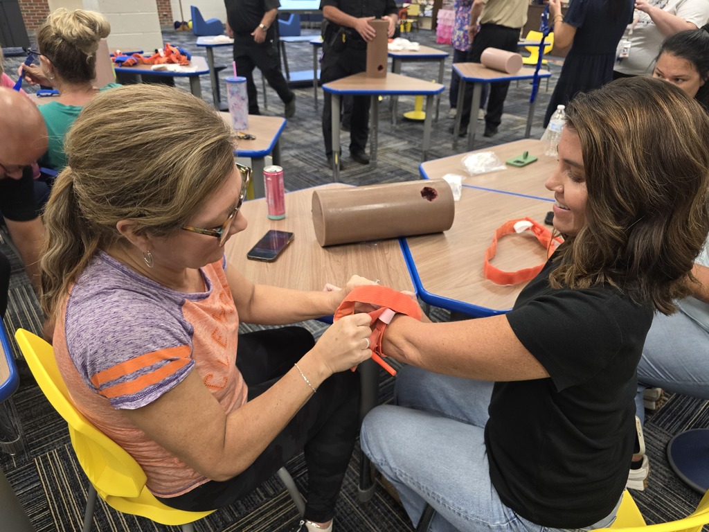 Two BMS staff members practice properly applying a tourniquet during hands-on Stop the Bleed training, with other staff and instructors participating in the background.