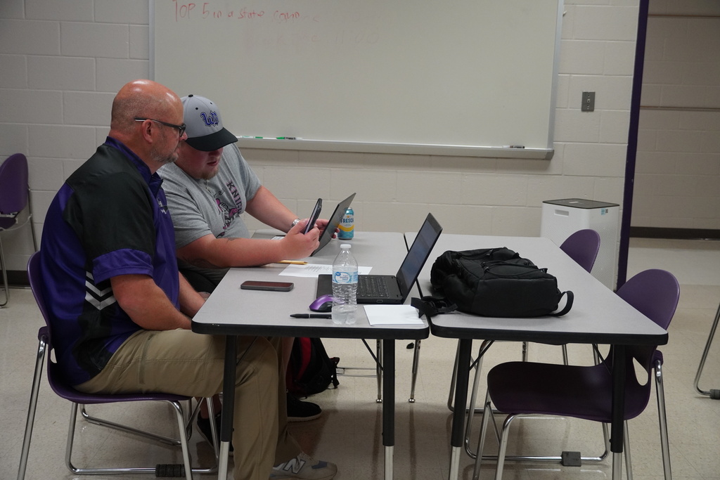 Two staff members sit in classroom desks working on laptops.