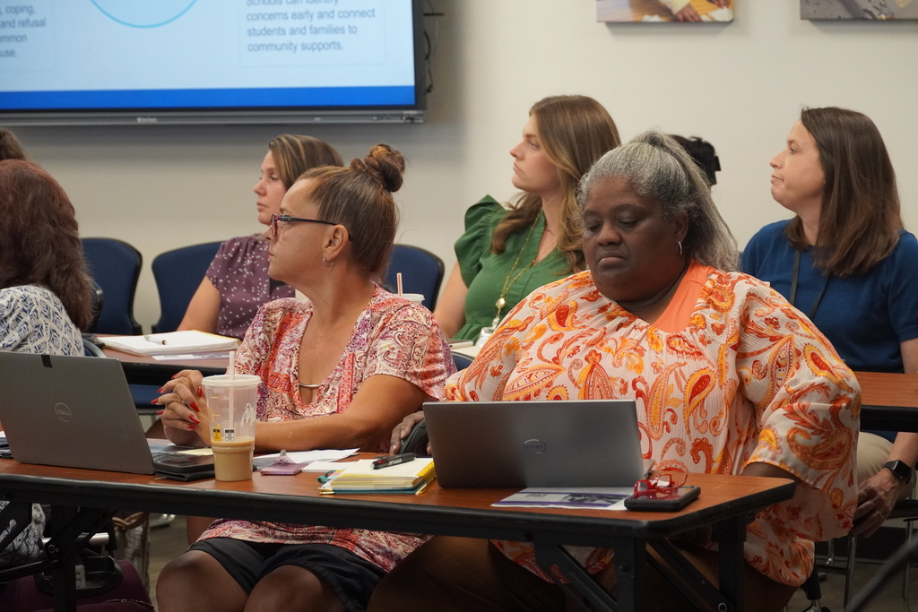 School counselors are sitting at long tables listening to a presentation.