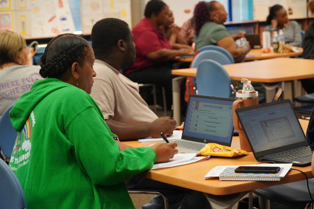Staff members are seen at tables with laptops open and taking notes.