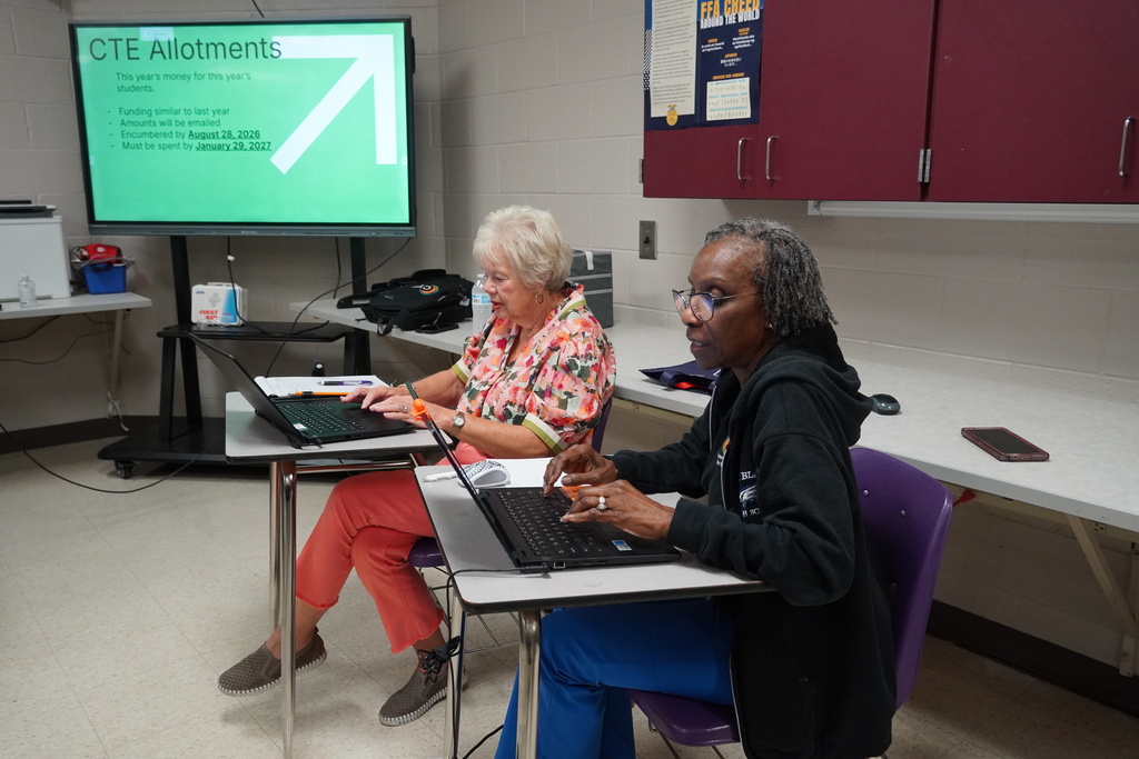 Two staff members sit in classroom desks working on laptops.