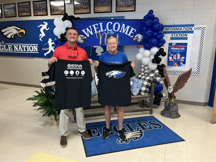 Man in red shirt, woman in blue shirt, holding up black T-shirts with eagles on them in front of a welcome backdrop with eagles, a bench, flowers, and balloons