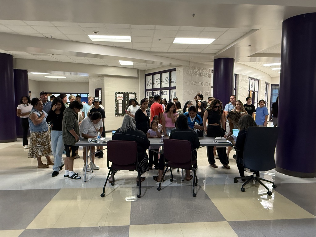 Students, parents, and staff gather around tables inside a bright school hallway during an orientation event.