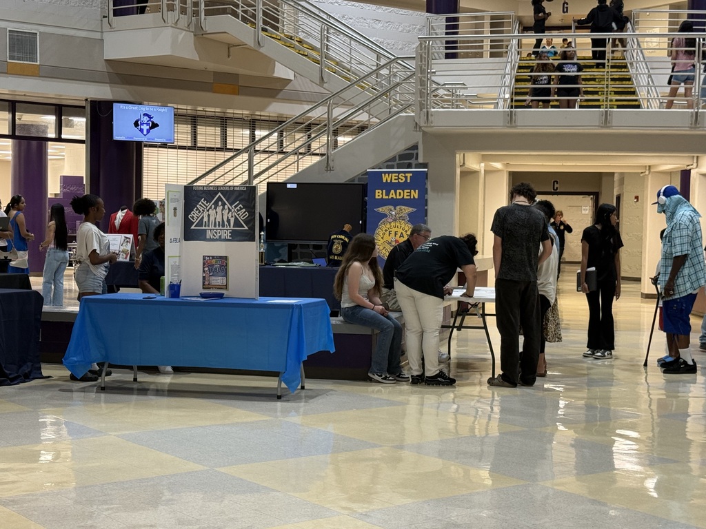 Students and families visit club tables, including FBLA and FFA, set up in a school lobby with stairs in the background.
