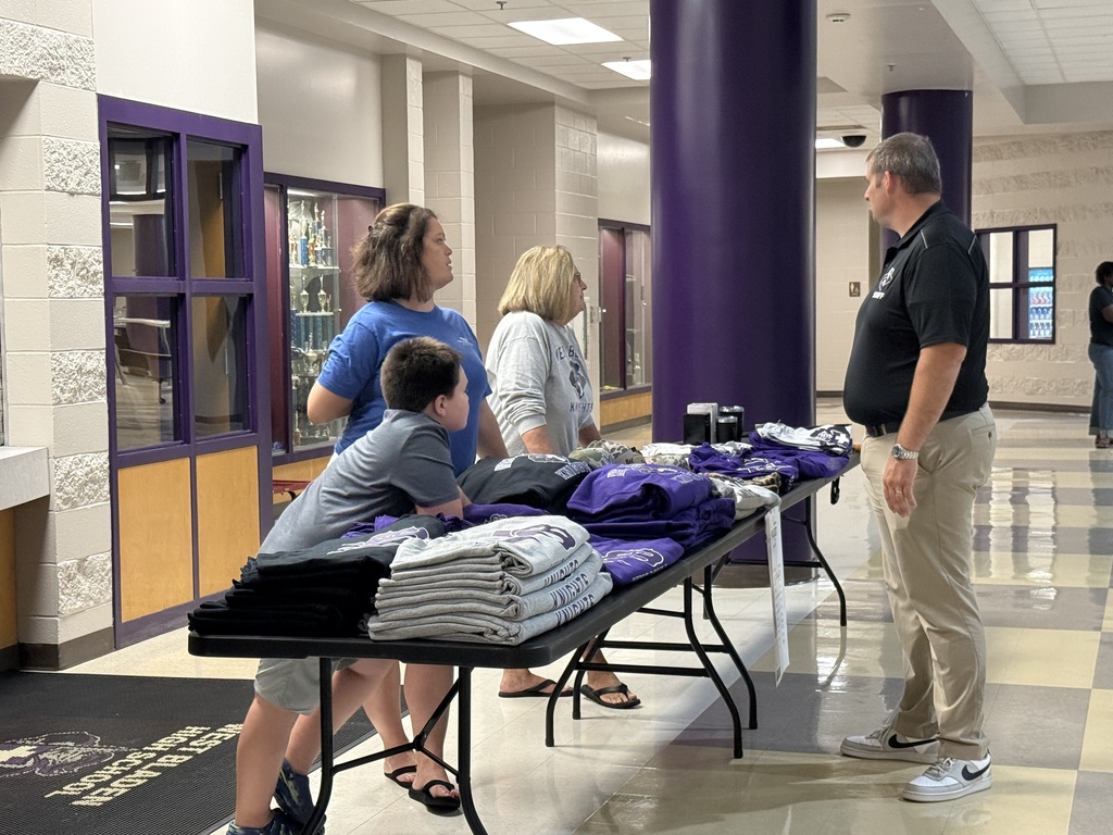 Students and parents browse school apparel stacked on a folding table while speaking with a staff member in a hallway.