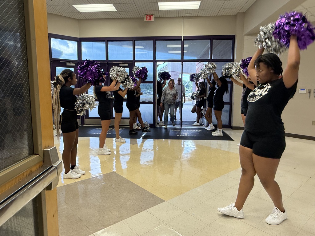 High school cheerleaders hold purple pom-poms and cheer inside a school entrance lobby to welcome arriving guests.