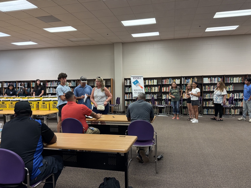 Students, parents, and staff gather in a school media center at informational tables during freshman orientation.
