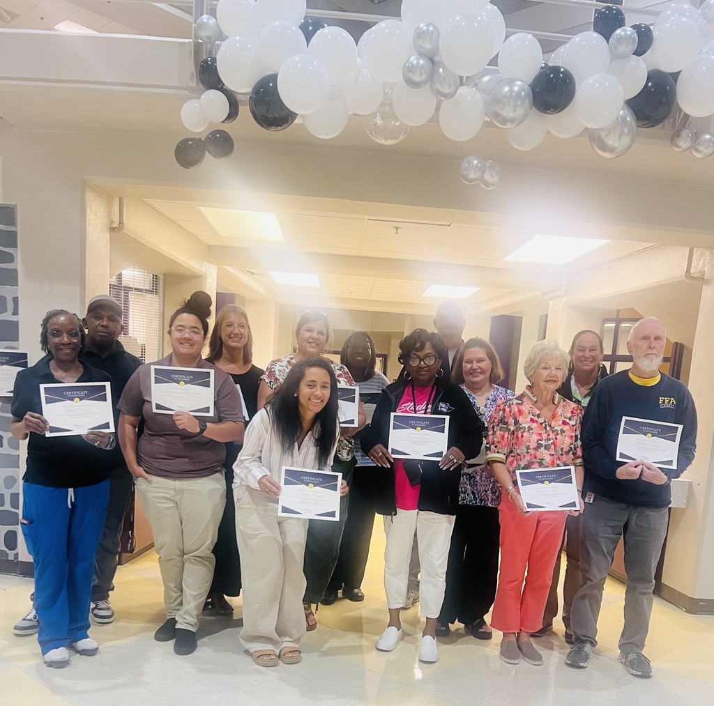 A diverse group of adults pose indoors beneath a large white, silver, and black balloon display. Several people in the front row hold certificates, smiling for a group photo at what appears to be an awards or recognition event.