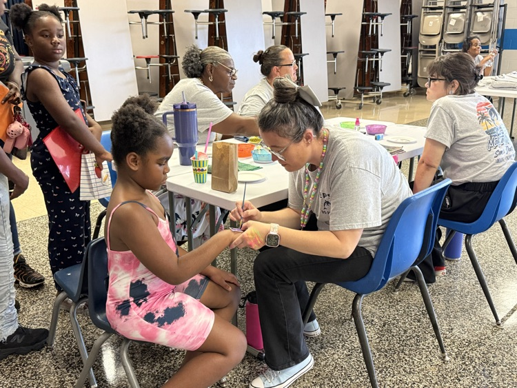     A staff member paints a colorful design on a student’s hand during Open House while families participate in activities nearby.