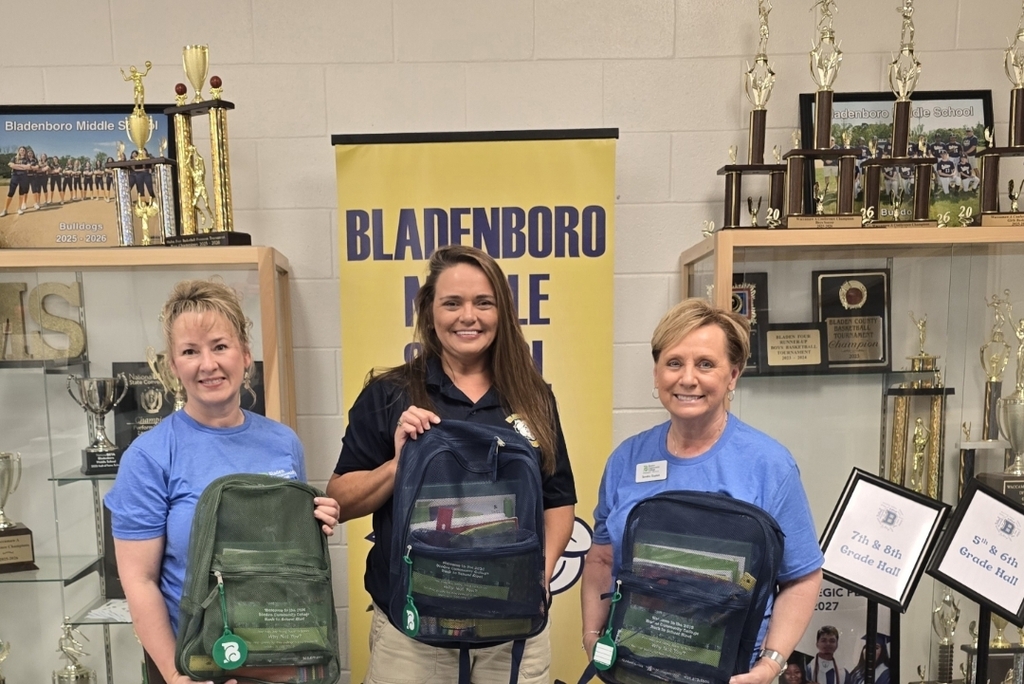 Three women stand together inside Bladenboro Middle School, smiling and holding backpacks filled with school supplies. A yellow and navy Bladenboro Middle School banner is displayed behind them, with trophy cases showcasing school awards and athletic achievements on both sides.