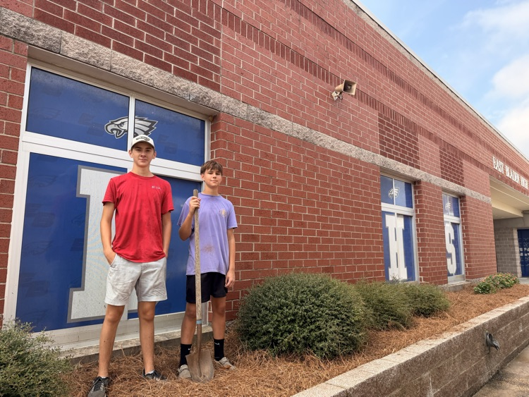 two guys win in a red shirt, khaki shorts, the other and purple shirt, black shorts, standing in a flower bed in front of a window with blue tint, holding a shovel