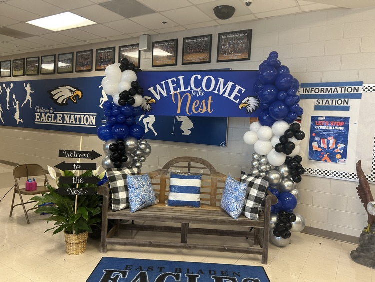 wooden bench with a plant beside it, balloon columns, and a banner across the top that Red is welcome and East Bladen eagle rug is in front of the bench and there is a eagle statue beside it