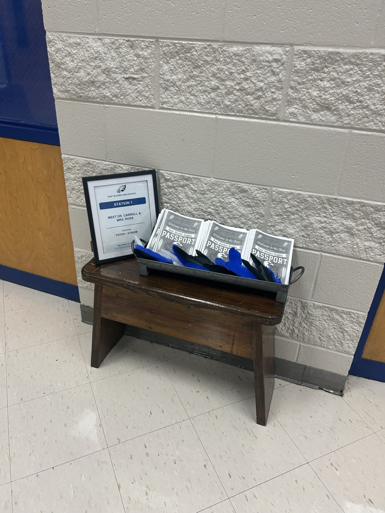 White tile floor white wall with a small wooden bench in front of it with a metal basket with East Bladen, passports in it and black and blue feathers