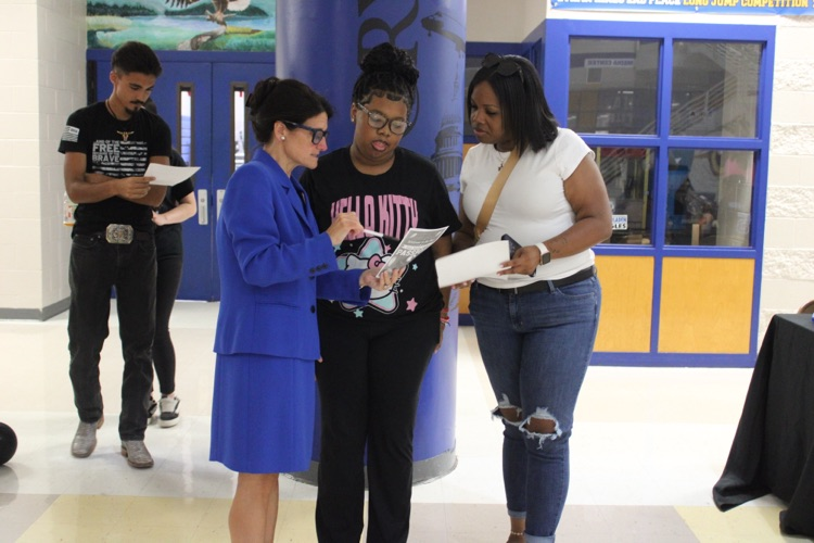 Lady in blue skirt suit stands explaining something to a student and black shirt and pants with a parent and jeans and a white shirt. They stand with a blue column behind them.