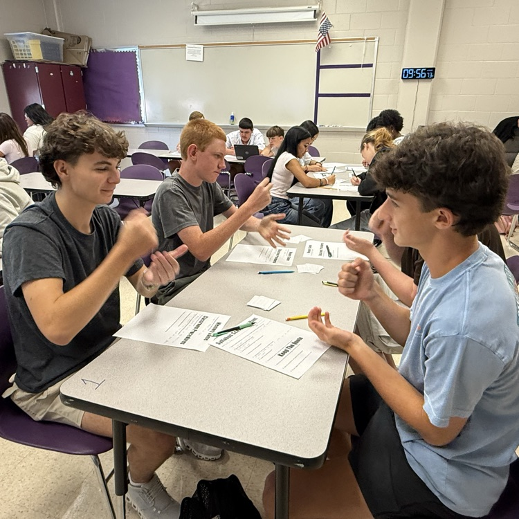 4 male students play rock paper scissor at one table  while other students work on a work sheet 