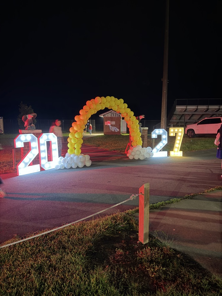 dark sky entrance to a football field with a yellow balloon arch with white cloud looking balloons at the bottom and the number is 20 and 27 on both side sides
