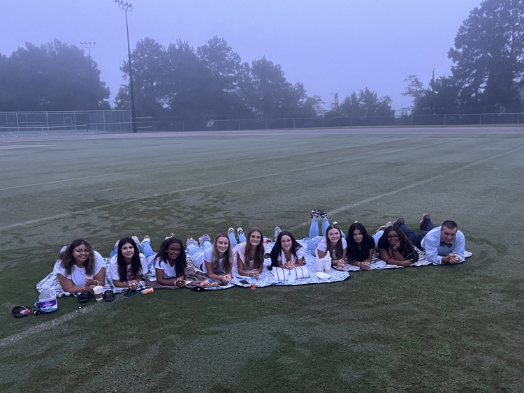 A group of 10 students and school principal lay smiling as a pose for a photo there is footprints all over the ground from the morning, dew and fog in the background