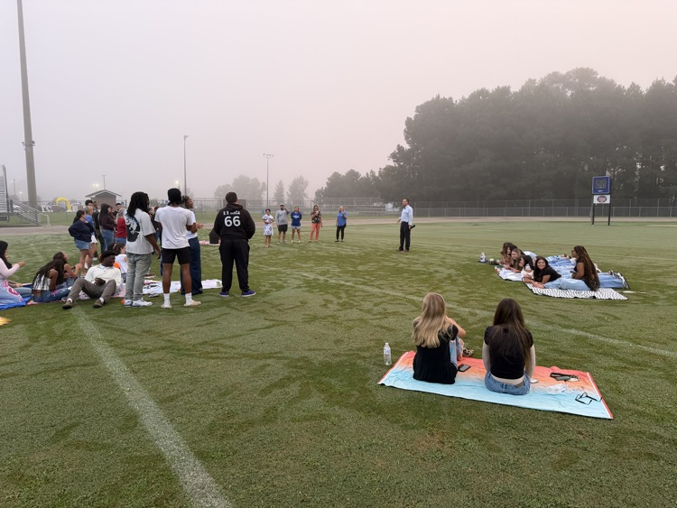 A large group of seniors and their parents are sitting and standing around a football field as the principal gives a motivational speech there is morning due on the football field and fog in the background